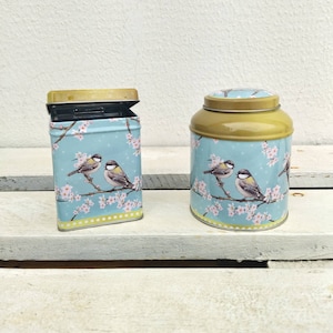 Könnte beinhalten: Zwei blaue und goldene Metall-Teedosen mit Blumenmuster und Vögeln. Die Dosen haben einen goldenen Deckel und einen blauen Boden mit einem weißen Blumenmuster und zwei Vögeln auf einem Ast.
