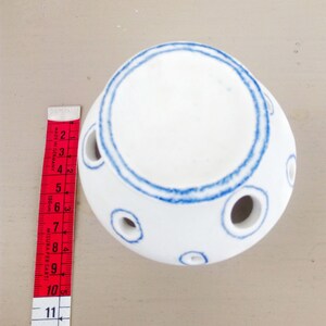 May include: A white ceramic sphere with blue hand-painted circular outlines around the top opening and several smaller circular openings along the sides. A red and white measuring tape is placed next to it, showing a height of 4 inches.