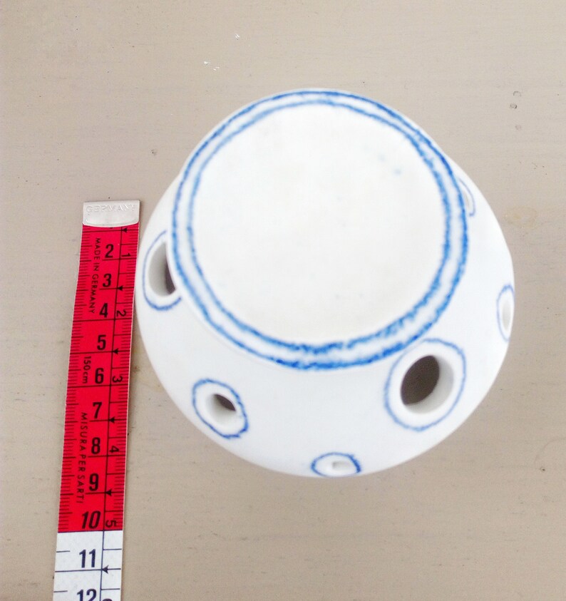 May include: A white ceramic sphere with blue hand-painted circular outlines around the top opening and several smaller circular openings along the sides. A red and white measuring tape is placed next to it, showing a height of 4 inches.