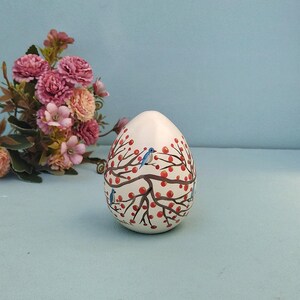 Può includere: Un ornamento liscio a forma di uovo color bianco sporco con un disegno dipinto a mano di rami d'albero marroni con piccole bacche rosse e diversi piccoli uccelli blu appollaiati sui rami che avvolgono l'intera superficie dell'oggetto.