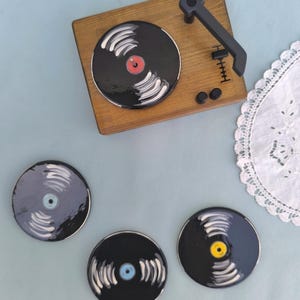 May include: A wooden turntable with a black vinyl record design, accompanied by three matching coasters. The coasters and the record on the turntable feature a black base with white grooves and colored center labels. The turntable has a black arm and buttons.