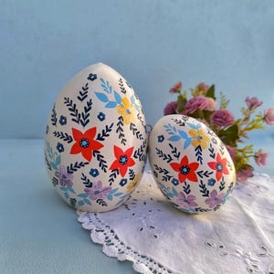 May include: Two egg-shaped decorative objects with a smooth cream-colored finish, hand-painted with a repeating pattern of red, yellow, and purple flowers, light blue leaves, and dark blue sprigs, arranged in a vertical orientation.