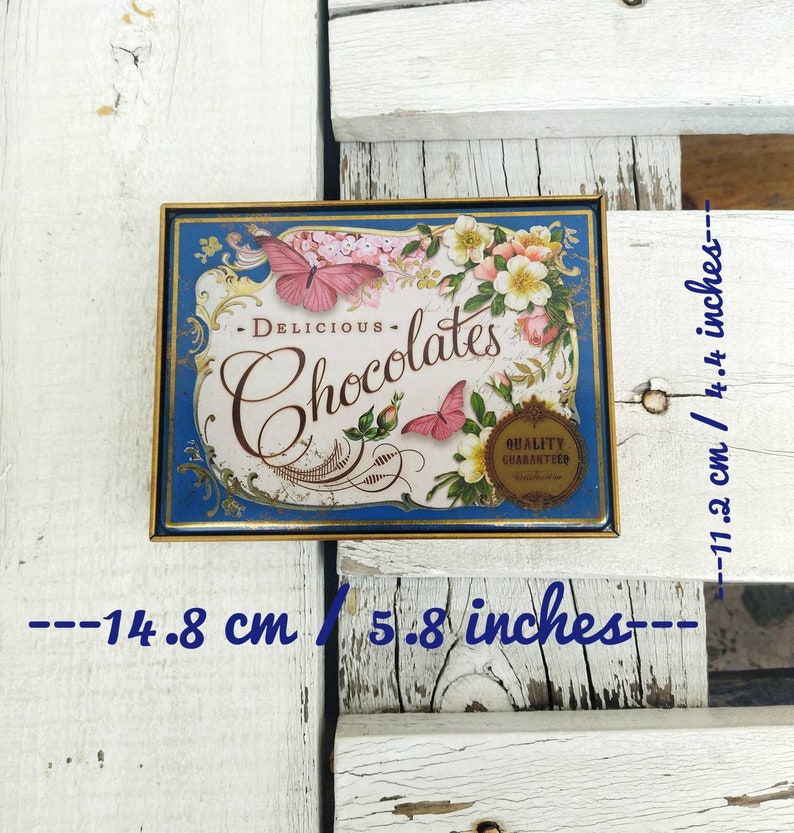 May include: A vintage tin box with a blue background and gold trim. The box features a floral design with butterflies and the text "DELICIOUS - Chocolates - QUALITY GUARANTEED". The box measures 5.8 inches by 4.4 inches.