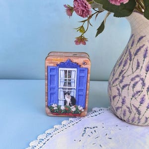 May include: A rectangular metal tin featuring a printed illustration of a brick wall with a white window, blue shutters, and a black and white cat sitting on the sill. A window box filled with white and red flowers sits below the window frame.