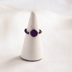 Könnte beinhalten: Ein schwarzer Ring mit einem runden schwarzen Stein und goldenen Perlen auf einem weißen, kegelförmigen Ringständer.