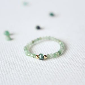 Puede incluir: Un delicado anillo hecho de pequeñas cuentas de piedras preciosas verdes con una cuenta de piedra preciosa verde más grande en el centro. El anillo está acentuado con dos pequeñas cuentas doradas a cada lado de la piedra central.