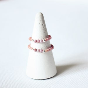 May include: Two delicate rings made of pink and white beads on a white cone-shaped ring holder.