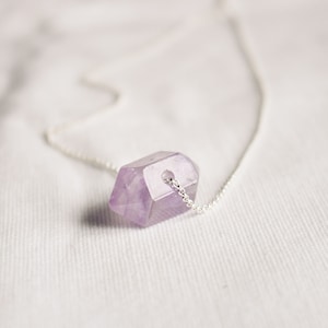 Könnte beinhalten: Eine zarte silberne Kettenkette mit einem einzelnen, facettierten Amethyst-Kristall-Anhänger. Der Kristall ist hellviolett und hat eine leicht unregelmäßige Form.