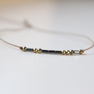 Puede incluir: Un brazalete delicado con un diseño simple que presenta cuentas negras y doradas en un cordón beige.