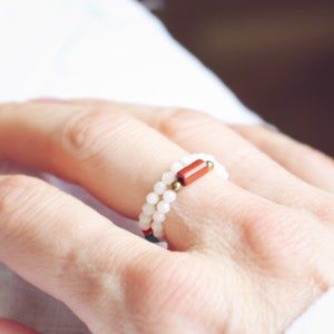 Könnte beinhalten: Ein weißer Perlenring mit einem roten und goldenen Akzent. Der Ring wird an einem Finger getragen.