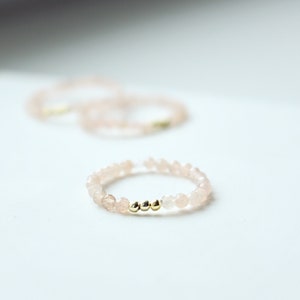 Könnte beinhalten: Ein zarter Ring aus kleinen rosa Perlen mit drei goldenen Perlen in der Mitte.