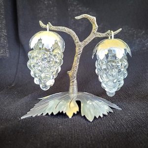 Op de afbeelding: Een zilverkleurige metalen standaard in de vorm van een boomtak met een bladvormige voet, die twee zout- en peperstrooiers van helder glas vasthoudt. Elke strooier is gevormd als een tros druiven met een zilverkleurige metalen dop aan de bovenkant.