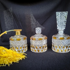May include: Three vintage glass perfume bottles with diamond-cut patterns and gold accents. One bottle has a yellow tassel and spray tube. The bottles are clear with yellow and gold details, set against a dark background.