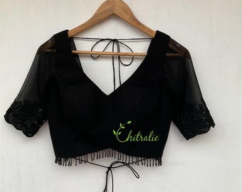 May include: Black blouse with a V-neck and sheer sleeves. The sleeves have floral lace detailing at the cuffs. The blouse has a beaded fringe along the bottom and the word "chitralie" in green script.