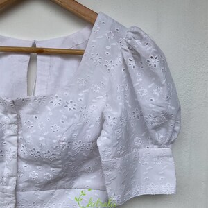 May include: White cotton blouse with a square neckline and puff sleeves. The blouse features a floral eyelet pattern and a button closure at the front.