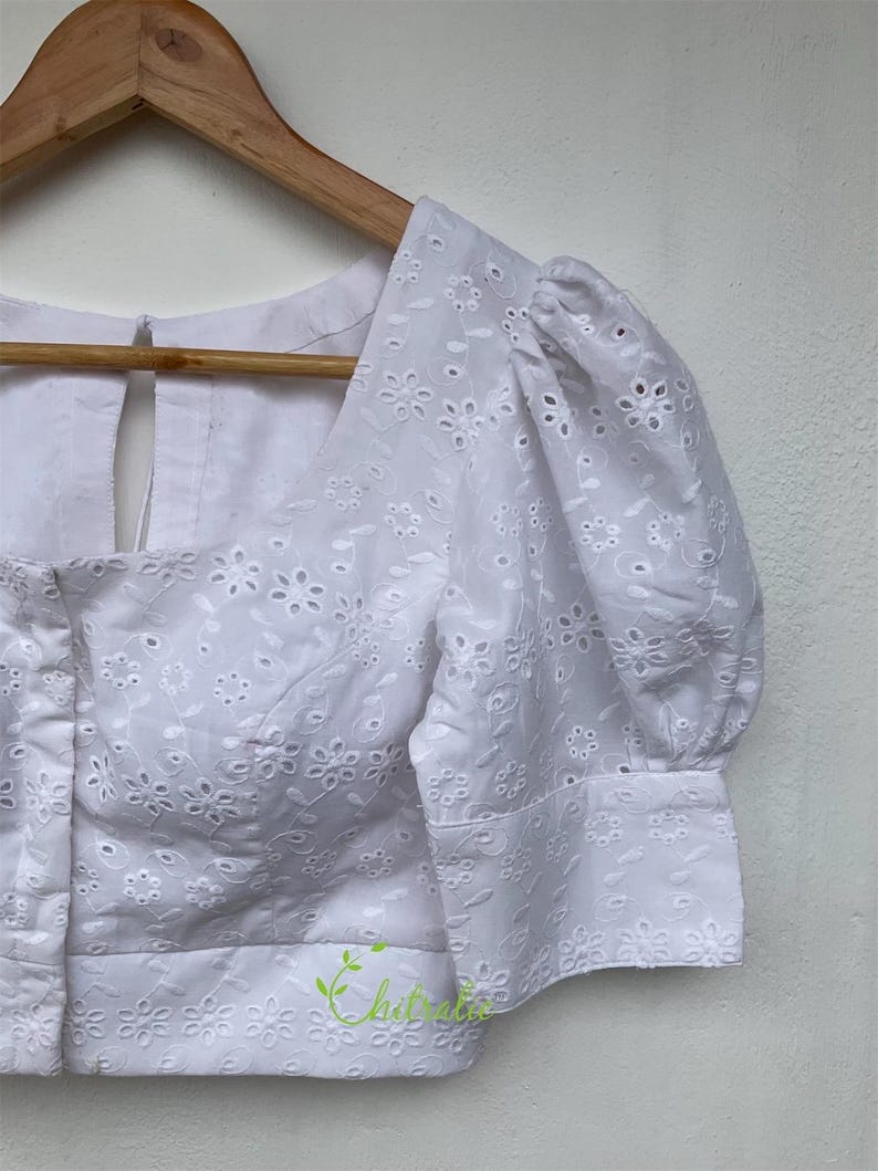 May include: White cotton blouse with a square neckline and puff sleeves. The blouse features a floral eyelet pattern and a button closure at the front.