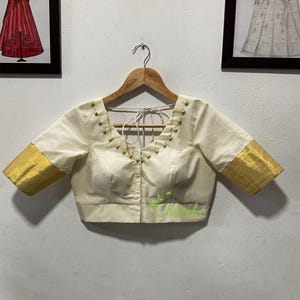 Peut inclure: Blouse de couleur crème avec des détails dorés. Le haut à manches courtes présente une bordure dorée sur les manches et de petites décorations dorées le long de l'encolure. Le chemisier est suspendu à un cintre en bois sur un fond neutre.