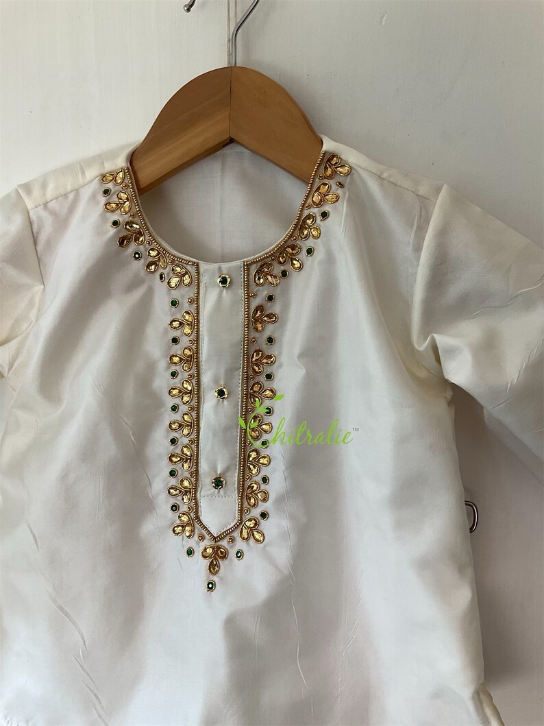 May include: White kurta with gold and green embroidery. The neckline and front of the garment are adorned with intricate embellishments.