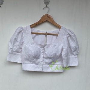 May include: White cotton blouse with a round neckline and short puffed sleeves. The blouse has a button-up front and is decorated with eyelet embroidery.
