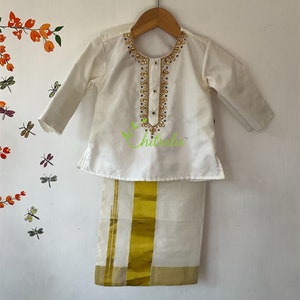 May include: White traditional Indian outfit for a child, featuring a long-sleeved shirt with gold and white embroidery and a white dhoti with a gold border.