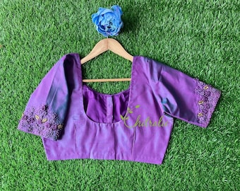 May include: A purple silk blouse with a scoop neck and short sleeves. The sleeves are decorated with purple and gold beading. The blouse is hanging on a wooden hanger against a green grass background.