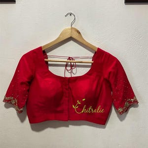 Könnte beinhalten: Eine leuchtend rote Bluse mit Ellenbogenärmeln, verziert mit goldener Stickerei an den Manschetten und Ärmeln. Die Bluse hat einen quadratischen Ausschnitt und das Wort "Chitralie" in Schreibschrift.