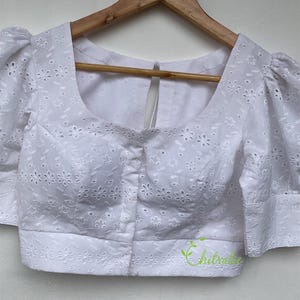 May include: White cotton blouse with a round neckline and short puffed sleeves. The blouse features a delicate eyelet pattern and a button closure at the front.
