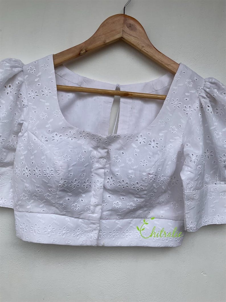May include: White cotton blouse with a round neckline and short puffed sleeves. The blouse features a delicate eyelet pattern and a button closure at the front.