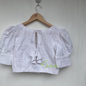 May include: White cotton blouse with a tie at the back and a floral pattern. The blouse has short, puffed sleeves.