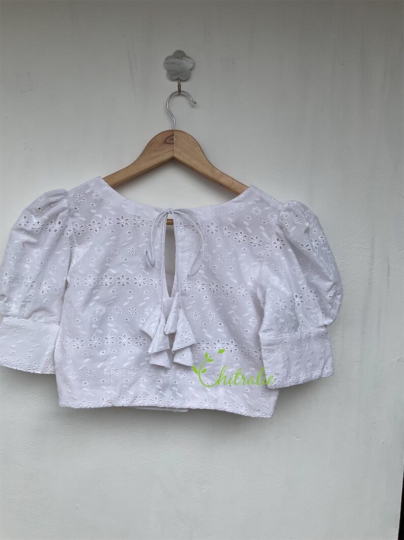 May include: White cotton blouse with a tie at the back and a floral pattern. The blouse has short, puffed sleeves.