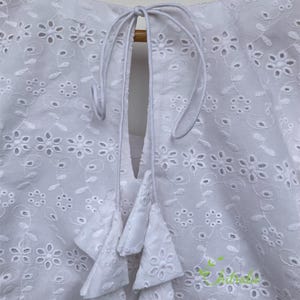May include: White cotton blouse with a round neckline and a tie at the back. The blouse features a delicate eyelet pattern and a tiered ruffle detail at the back.