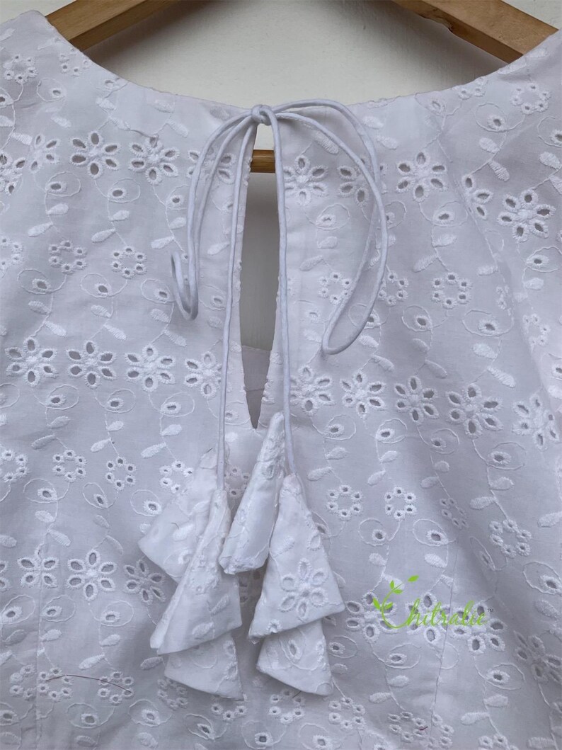 May include: White cotton blouse with a round neckline and a tie at the back. The blouse features a delicate eyelet pattern and a tiered ruffle detail at the back.