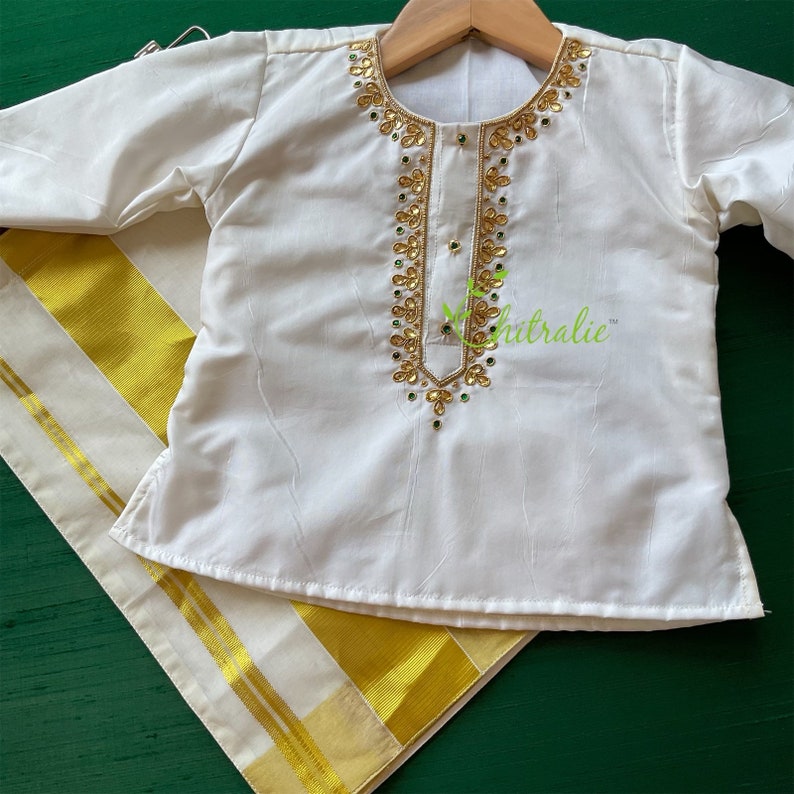 May include: White traditional Indian outfit for a child, featuring a long-sleeved shirt with gold and green embroidery and a white and gold dhoti.