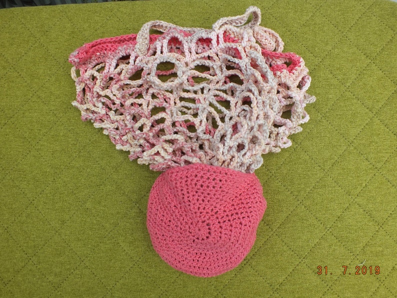 May include: A pink and white crocheted mesh bag with a pink crocheted hat. The bag is lying on a green surface.