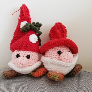 Könnte beinhalten: Zwei gehäkelte Gnome mit roten Mützen und rosafarbenen Gesichtern. Ein Gnom hat einen grünen, blättrigen Schmuck an seiner Mütze. Die Gnome sitzen auf einer grauen, strukturierten Oberfläche.