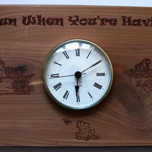 Puede incluir: Reloj de madera con esfera redonda y números romanos. El reloj está incrustado en una tabla de madera rectangular con ilustraciones de ranas grabadas y el texto "Time's Fun When You're Having Flies!"