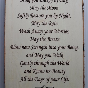 May include: A wooden sign with a white background and brown text. The sign features a poem about the sun, moon, rain, and breeze. The poem reads: "May the Sun Bring you Energy by Day, May the Moon Softly Restore you by Night, May the Rain Wash Away your Worries, May the Breeze Blow new Strength into your Being, and May you Walk Gently through the World and Know its Beauty All the Days of your Life."