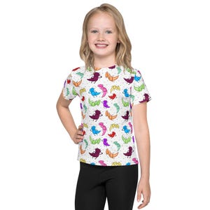 Könnte beinhalten: Ein weißes T-Shirt mit einem sich wiederholenden Muster aus bunten Cartoon-Kreaturen, darunter Lila, Blau, Orange und Grün. Kleine Polka Dots sind über den Hintergrund verstreut. Das kurzärmelige T-Shirt wird getragen.