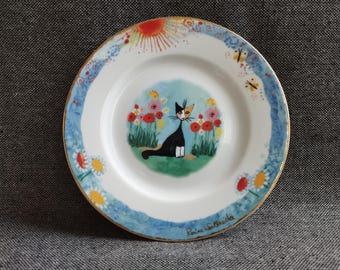 May include: A white porcelain plate with a blue and gold rim. The plate features a black cat with white markings sitting in a field of flowers. The cat is looking to the right. The plate has a sun and butterflies on the rim. The text "Romina Laetitia" is written on the bottom of the plate.