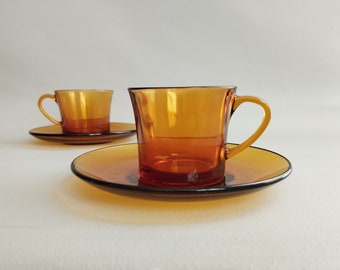 May include: Two amber glass coffee cups with matching saucers. The cups have a rounded shape and a flared lip. The saucers are slightly larger than the cups and have a smooth, rounded edge.