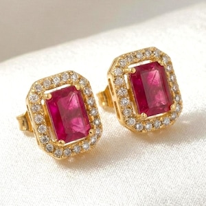 May include: A pair of gold stud earrings featuring rectangular, deep red gemstones. Each earring is encircled by a halo of small, clear gemstones. The earrings are set against a white, textured background.