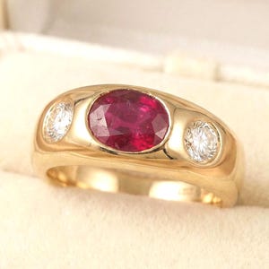 Puede incluir: Un anillo de oro con una gran piedra central ovalada roja y dos piedras blancas más pequeñas a cada lado. El anillo está engastado en una banda de oro pulido y se muestra en una caja blanca.
