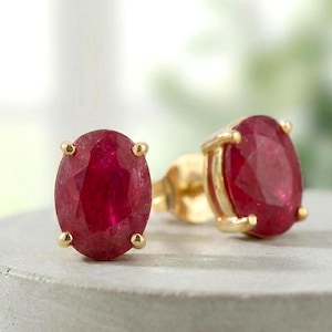 Pode incluir: Um par de brincos de rubi ovais com engastes dourados. Os rubis vermelho-escuros são definidos em uma configuração de quatro garras. Os brincos são exibidos em uma superfície cinza.