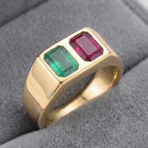 Puede incluir: Un anillo de oro con dos piedras preciosas rectangulares: una esmeralda verde y un rubí rojo. El anillo tiene un acabado pulido y un diseño clásico. Las piedras están colocadas una al lado de la otra en un engaste rectangular.