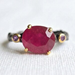 Peut inclure: Gros plan sur une bague avec un grand rubis facetté rouge foncé en son centre. Le rubis est maintenu par quatre griffes dorées. L'anneau est gris foncé avec deux petites pierres violettes de chaque côté.