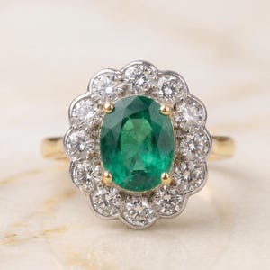 May include: A ring featuring a central oval-cut green emerald held by four yellow gold prongs, surrounded by a scalloped halo of twelve round brilliant-cut white diamonds set in white metal, mounted on a polished yellow gold band.