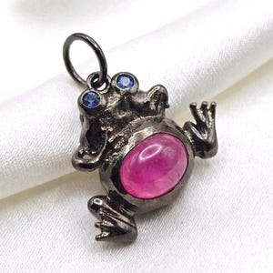 Puede incluir: Colgante de rana de metal oscuro con una gran gema rosa ovalada en el vientre y dos pequeñas gemas azules para los ojos. La rana está detallada con piel texturizada y extremidades extendidas.