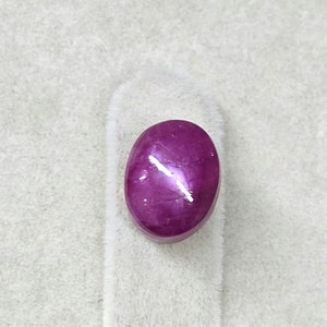 May include: A polished, oval-shaped ruby gemstone. The gem is a deep, saturated pinkish-red color with a glossy surface. It is set on a white, fabric-covered display stand, showcasing its shape and color.