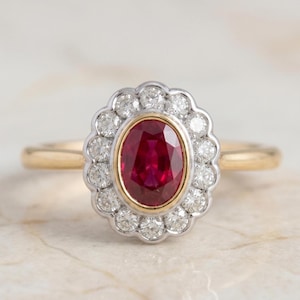 Könnte beinhalten: Ein goldener Ring mit einem ovalen, tiefroten Rubin in der Mitte, umgeben von einem Kranz aus kleinen, runden Diamanten. Der Rubin ist tiefrot, und die Fassung ist goldfarben. Der Ring hat ein goldenes Band.