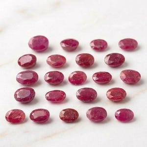 May include: A collection of oval-cut, deep red gemstones, possibly rubies, arranged on a white surface. The gems vary slightly in shade and size, with a polished, reflective surface. The arrangement suggests a display of loose stones.